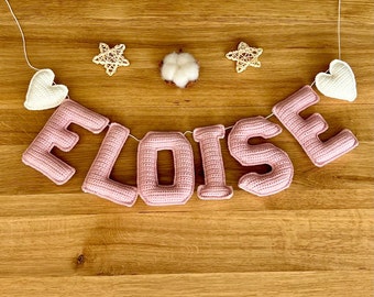 May include: A pink crocheted banner spelling "ELOISE" hangs against a wooden background. The banner includes white crocheted hearts and star-shaped decorations. A cotton ball is centered above the banner.