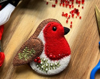 May include: A felt robin ornament featuring a bright red chest, brown wings, and a white belly. The wing is decorated with green embroidered sprigs, while the chest is accented with small red beads and white bead clusters near the bottom edge.