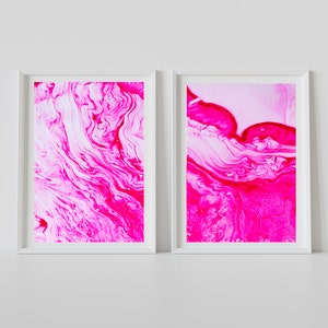 K&ouml;nnte beinhalten: Zwei gerahmte abstrakte Kunstdrucke mit wirbelnden Mustern in Pink und Wei&szlig;. Die Drucke sind in wei&szlig;en Rahmen und h&auml;ngen an einer wei&szlig;en Wand.