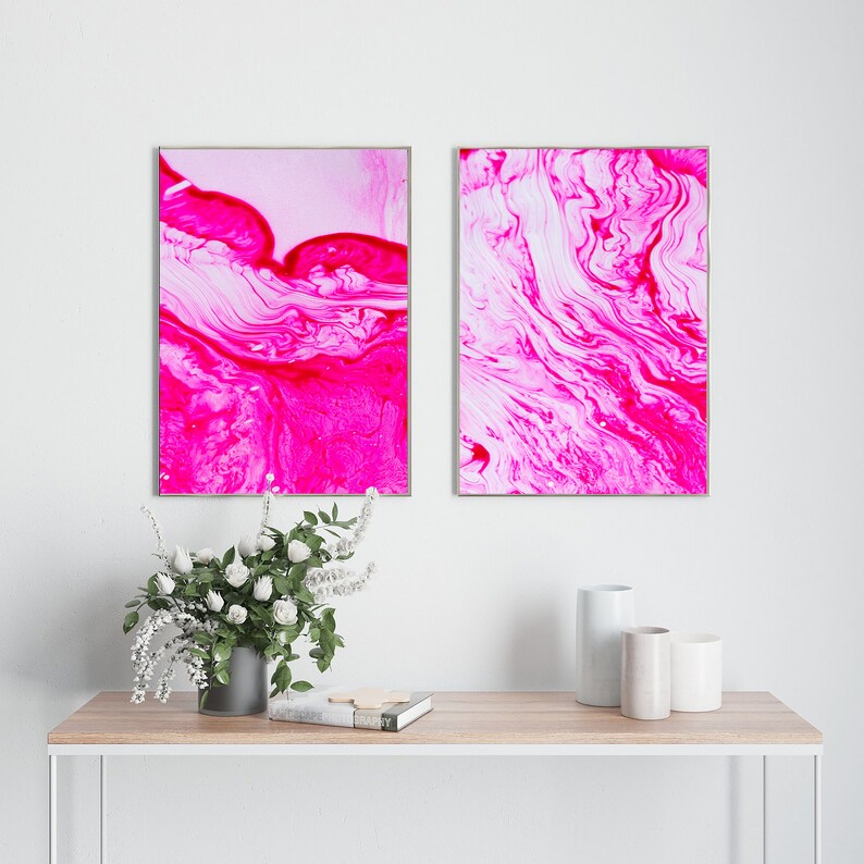 K&ouml;nnte beinhalten: Zwei gerahmte abstrakte Gem&auml;lde mit pinken und wei&szlig;en wirbelnden Mustern. Die Gem&auml;lde h&auml;ngen an einer wei&szlig;en Wand &uuml;ber einem Holztisch mit einer Vase mit wei&szlig;en Blumen und drei dekorativen Zylindern.