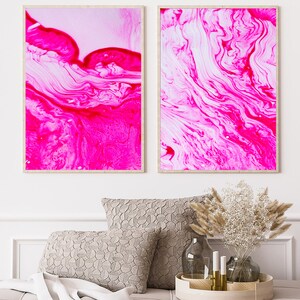 K&ouml;nnte beinhalten: Zwei gerahmte abstrakte Kunstdrucke mit wirbelnden Mustern in Pink und Wei&szlig;. Die Drucke h&auml;ngen an einer wei&szlig;en Wand &uuml;ber einem Sofa mit Kissen und einem dekorativen Tablett.