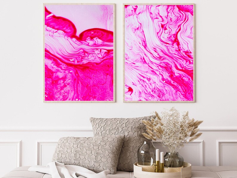 K&ouml;nnte beinhalten: Zwei gerahmte abstrakte Kunstdrucke mit wirbelnden Mustern in Pink und Wei&szlig;. Die Drucke h&auml;ngen an einer wei&szlig;en Wand &uuml;ber einem Sofa mit Kissen und einem dekorativen Tablett.