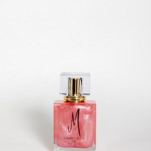 May include: A square, clear glass bottle filled with pink shimmer body oil. The bottle has a gold-colored neck and a clear, rectangular cap. The label features a black "M" and the words "SHIMMER BODY OIL" and "50 ML / 1.7 FL OZ."