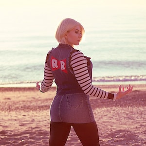 May include: A woman wearing a blue denim vest with a red "RR" patch, a striped shirt, black leggings, and brown boots. She is standing on a sandy beach with the ocean in the background.