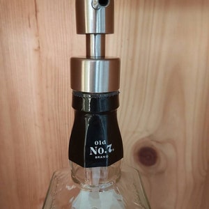 May include: A clear glass bottle with a black label that reads "old No. 7. BRAND" and a silver soap dispenser pump on top. The bottle is sitting on a wooden surface.