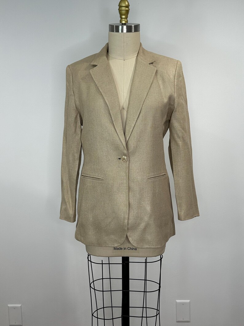 K&ouml;nnte beinhalten: Ein beigefarbener Blazer mit einem Ein-Knopf-Verschluss und zwei Vordertaschen. Der Blazer wird auf einer Schaufensterpuppe pr&auml;sentiert. Auf dem Etikett der Schaufensterpuppe steht "Made in China".