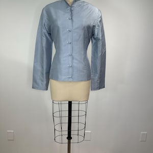 以下が含まれることがあります： マンダリンカラーとボタン留めのフロントが付いた、水色の長袖ジャケット。マネキンに展示されています。生地は微妙な光沢があり、シルクまたはサテンを示唆しています。ジャケットのデザインはシンプルでエレガントです。