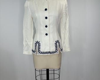 Puede incluir: Chaqueta vintage color crema con mangas largas y cuello redondo. La chaqueta presenta botones azul marino y bordados decorativos en el dobladillo. La chaqueta se exhibe en un maniquí.