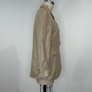 K&ouml;nnte beinhalten: Ein beigefarbener Blazer auf einer Schaufensterpuppe. Der Blazer hat ein fallendes Revers, zwei Vordertaschen und drei Kn&ouml;pfe an jeder Manschette. Der Stoff scheint strukturiertes Leinen oder eine Leinenmischung zu sein. Der Hals der Schaufensterpuppe ist mit der Nummer 12 markiert.