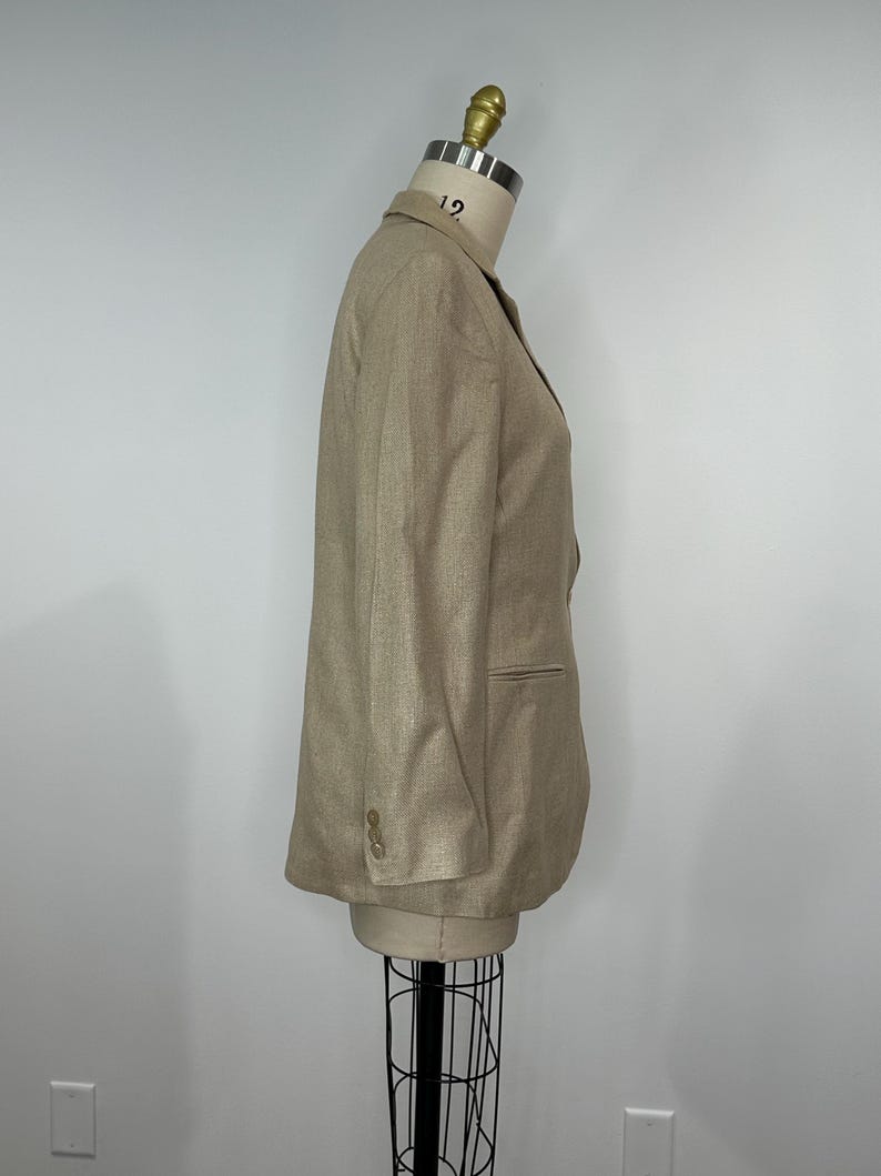 K&ouml;nnte beinhalten: Ein beigefarbener Blazer auf einer Schaufensterpuppe. Der Blazer hat ein fallendes Revers, zwei Vordertaschen und drei Kn&ouml;pfe an jeder Manschette. Der Stoff scheint strukturiertes Leinen oder eine Leinenmischung zu sein. Der Hals der Schaufensterpuppe ist mit der Nummer 12 markiert.