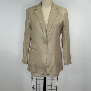K&ouml;nnte beinhalten: Ein beigefarbener Blazer mit einem Ein-Knopf-Verschluss, zwei Vordertaschen und einem Revers. Der Blazer wird auf einer Schaufensterpuppe pr&auml;sentiert. Das Etikett auf der Schaufensterpuppe lautet "Made in China".