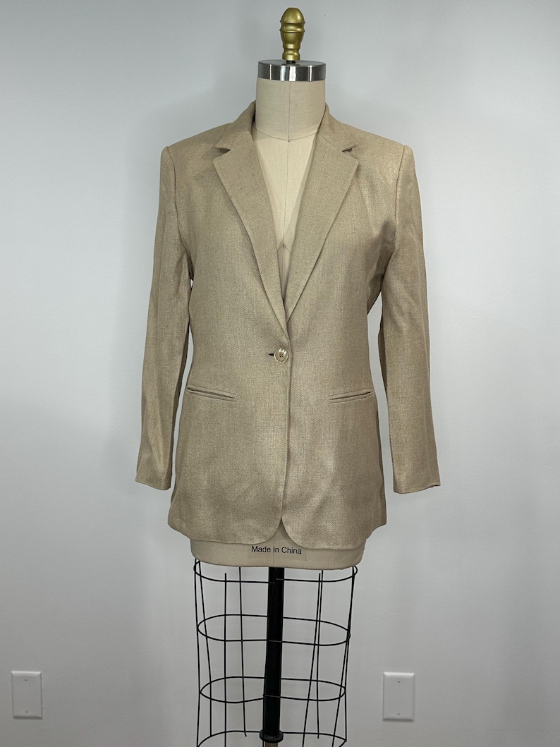 K&ouml;nnte beinhalten: Ein beigefarbener Blazer mit einem Ein-Knopf-Verschluss, zwei Vordertaschen und einem Revers. Der Blazer wird auf einer Schaufensterpuppe pr&auml;sentiert. Das Etikett auf der Schaufensterpuppe lautet "Made in China".