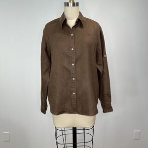 May include: A brown button-down shirt with long sleeves and a collar. The shirt has white buttons and a small logo on the chest. The sleeves can be rolled up and secured with a button.