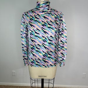 Könnte beinhalten: Ein langärmliges Rollkragentop mit einem farbenfrohen, abstrakten Muster. Das Oberteil hat einen Mix aus rosa, blauen, gelben und grünen Streifen auf hellem Hintergrund. Das Kleidungsstück wird auf einer Schaufensterpuppe präsentiert.