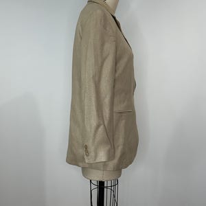K&ouml;nnte beinhalten: Ein beigefarbener Blazer, der auf einer Schaufensterpuppe pr&auml;sentiert wird. Der Blazer hat ein fallendes Revers, zwei Kn&ouml;pfe und einreihiges Design. Die &Auml;rmel haben drei Kn&ouml;pfe am B&uuml;ndchen. Der Blazer ist Gr&ouml;&szlig;e 12.
