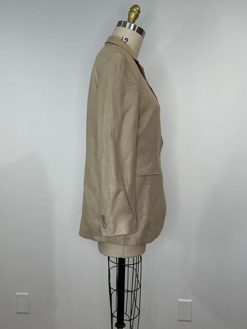 K&ouml;nnte beinhalten: Ein beigefarbener Blazer, der auf einer Schaufensterpuppe pr&auml;sentiert wird. Der Blazer hat ein fallendes Revers, zwei Kn&ouml;pfe und einreihiges Design. Die &Auml;rmel haben drei Kn&ouml;pfe am B&uuml;ndchen. Der Blazer ist Gr&ouml;&szlig;e 12.