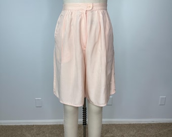 Puede incluir: Pantalones cortos de cintura alta de color rosa pálido con cierre de botón y pliegues. Los pantalones cortos se exhiben en un maniquí, mostrando su diseño a medida y su color claro. Los pantalones cortos tienen un estilo clásico y vintage.