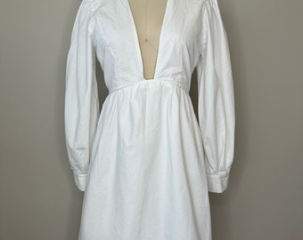 May include: A white mini dress with a deep V-neckline and long sleeves. The dress has a fitted bodice and a flowy skirt.
