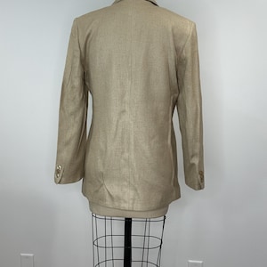 K&ouml;nnte beinhalten: Ein beigefarbener Blazer mit langen &Auml;rmeln und goldenen Kn&ouml;pfen an den &Auml;rmeln. Der Blazer wird auf einer Schaufensterpuppe pr&auml;sentiert. Der Stoff hat eine subtile Textur. Der Blazer ist eine neutrale Farbe, passend f&uuml;r verschiedene Outfits.