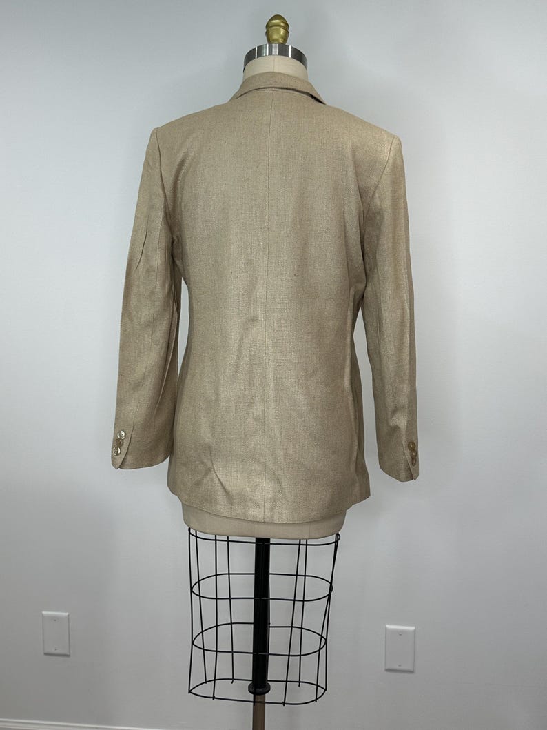 K&ouml;nnte beinhalten: Ein beigefarbener Blazer mit langen &Auml;rmeln und goldenen Kn&ouml;pfen an den &Auml;rmeln. Der Blazer wird auf einer Schaufensterpuppe pr&auml;sentiert. Der Stoff hat eine subtile Textur. Der Blazer ist eine neutrale Farbe, passend f&uuml;r verschiedene Outfits.