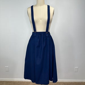 May include: A navy blue skirt with suspenders is displayed on a mannequin. The skirt is gathered at the waist and flares out. The suspenders are attached with gold-colored buttons. The skirt is knee-length.