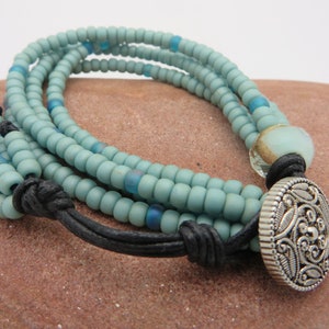 以下が含まれることがあります： ライトブルーのビーズが連なったマルチストランドブレスレット。青とクリアのビーズ、黒革コード、装飾的なシルバーカラーのボタン留めがアクセント。ユニークなジュエリーです。