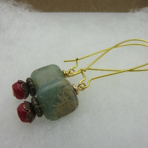 May include: A pair of handmade earrings featuring gold-colored ear wires. Each earring has a light green, square-shaped bead, a red bead, and decorative bronze-colored accents. The earrings are displayed on a white surface.