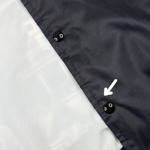 May include: Close-up of a black jacket with two black snap buttons. The jacket has a white lining and a dark gray exterior. A white arrow points to the snap buttons.