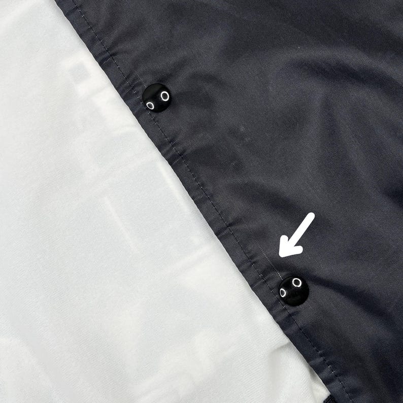 May include: Close-up of a black jacket with two black snap buttons. The jacket has a white lining and a dark gray exterior. A white arrow points to the snap buttons.