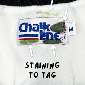 May include: Close-up of a white garment with a black collar and a tag that reads "Chalk Line" in blue lettering. The tag has red and green stripes. The text "STAINING TO TAG" is below the tag. There are stains on the tag.