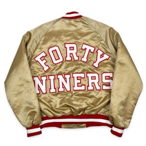 May include: A gold satin bomber jacket featuring the text FORTY NINERS in large white letters with red outlines across the back. The jacket includes a ribbed collar, cuffs, and hem with red and white stripes, and a snap button front closure.