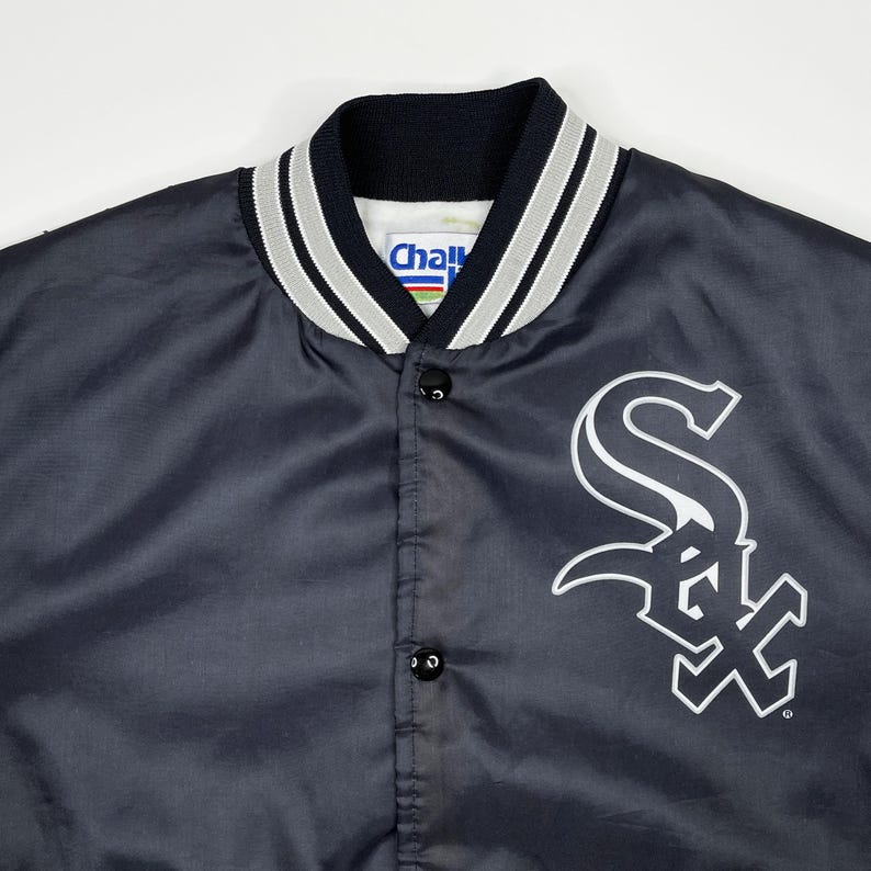 May include: A navy blue satin jacket with the Chicago White Sox logo in white on the front. The jacket has a black collar with white and gray stripes and a snap-button closure. The tag reads "Chall".