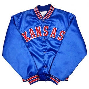 May include: A blue satin jacket with the word "KANSAS" in red block letters across the chest. The jacket has a button-up front, a ribbed collar, and cuffs with red and white stripes. The jacket is a medium size.