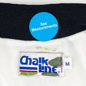 May include: Close-up of a white garment with a black collar. The garment has a tag that reads "Chalk Line" in blue and white. The tag also has a red and green stripe. A blue circle with the text "See Measurements" is visible.