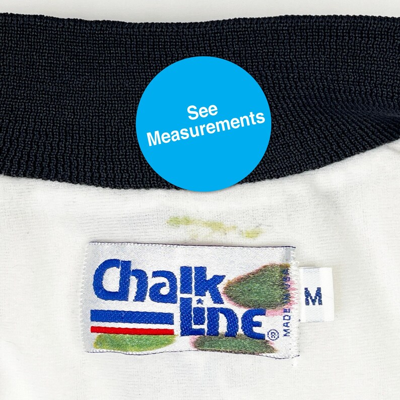 May include: Close-up of a white garment with a black collar. The garment has a tag that reads "Chalk Line" in blue and white. The tag also has a red and green stripe. A blue circle with the text "See Measurements" is visible.
