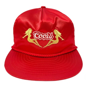 May include: A red satin baseball cap with a curved brim. The cap features the word "Coors" in white script, flanked by two gold-colored silhouette figures. A braided rope detail accents the brim.