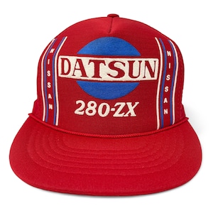 May include: Red baseball cap with a flat peak. The cap displays the word "DATSUN" in a white rectangle, a blue circle, and the text "280-ZX". The sides have vertical stripes with the word "NISSAN".