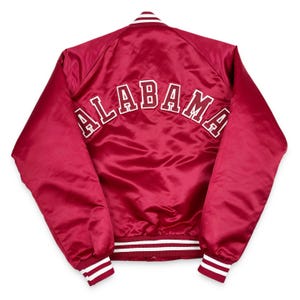 May include: A crimson satin bomber jacket with the word "ALABAMA" arched across the back in white block letters. The jacket has white and red striped cuffs, collar, and hem. This jacket is a piece of collegiate apparel.