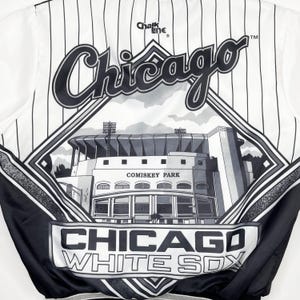 May include: White and black Chicago White Sox satin jacket with pinstripes. Features the word "Chicago" in a cursive font, a diamond-shaped graphic of Comiskey Park, and the words "Chicago White Sox". The jacket is from Chalk Line.