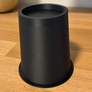 May include: A black, cylindrical object with a wide base and a slightly tapered body. The top has a recessed area. The item is made of a matte material and sits on a wooden surface. The object appears to be a furniture riser.