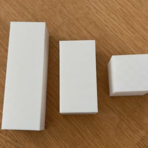 以下が含まれることがあります： 木製の表面に配置された、さまざまなサイズの3つの白い長方形のブロック。 ブロックは無地で、すっきりとしたミニマリストな外観です。 最大のブロックは高く細く、最小のブロックは立方体です。