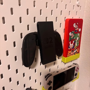 May include: A white pegboard with various gaming accessories attached. A black Nintendo Switch controller and charging dock are mounted, along with a red case featuring a Mario design. Additional game consoles are also displayed.