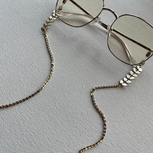 Könnte beinhalten: Goldene Ketten-Brillenhalterung mit weißen, blattförmigen Akzenten. Die Kette ist mit kleinen goldenen Schlaufen an der Brille befestigt.