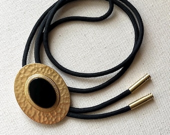 Puede incluir: Una corbata bolo con un cordón negro y puntas de metal dorado. La pieza central es un adorno ovalado de tono dorado con textura martillada, que rodea una incrustación negra lisa y ovalada enmarcada por un borde dorado con cuentas.