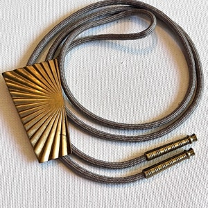 Puede incluir: Un bolo tie dorado con un diseño de rayos de sol y un cordón gris. El cordón tiene dos puntas de latón.
