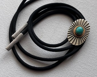 May include: A black bolo tie with a silver and gold oval pendant featuring a turquoise stone. The pendant has a sunburst design.