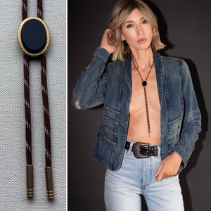 May include: A black onyx bolo tie with a gold-toned setting. The bolo tie is worn by a person with long blonde hair, wearing a denim jacket and jeans.