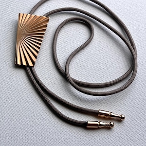Könnte beinhalten: Eine bronzefarbene Bolo-Krawatte mit einem geometrischen Anhänger im Sonnenstrahlen-Design. Der Anhänger ist rechteckig mit einem fächerartigen Muster. Das Band ist dunkelgrau, und die Spitzen sind bronzefarben mit kleinen Kugelenden. Gezeigt auf einer hellgrauen Oberfläche.