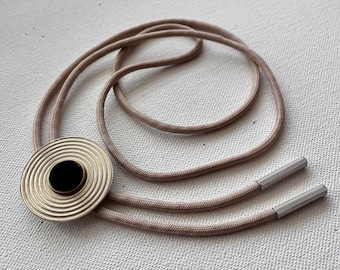 May include: A beige bolo tie with a gold and black circular pendant. The pendant has a concentric circle design with a black center.