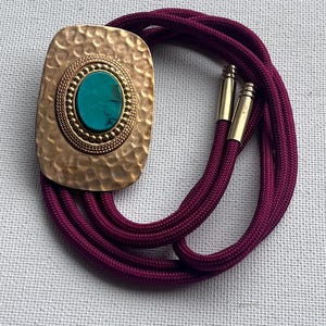 May include: A bolo tie featuring a hammered gold-tone rectangular pendant with a turquoise stone center. The pendant is attached to a deep purple cord with gold-tone metal tips. The bolo tie is a vintage accessory.